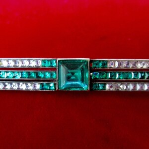 May include: A silver bar brooch with three rows of emerald green and clear rhinestones. The center stone is a larger square emerald green rhinestone.