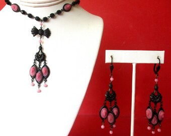May include: A black necklace with a pink cameo pendant and matching earrings. The necklace has a black bow at the top and the pendant has three pink cameos. The earrings have three pink cameos each.