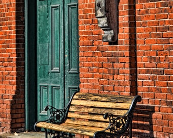 May include: A wooden bench with black cast iron supports sits in front of a red brick wall and a green door. The bench has a slatted seat and ornate armrests. The scene is bathed in sunlight, casting shadows on the wall.