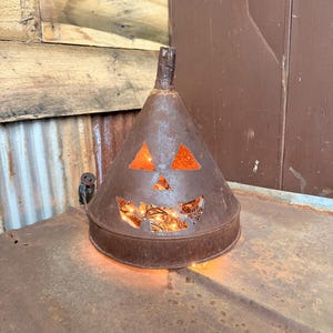 May include: A rustic, metal Halloween decoration shaped like a jack-o'-lantern. The brown, cone-shaped piece has cut-out eyes and a mouth, with orange accents. The interior glows with warm, yellow lights, creating a festive ambiance. The piece is sitting on a metal surface.