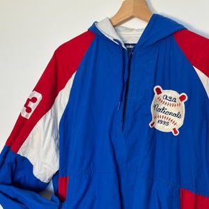 May include: A blue, red and white windbreaker jacket with a zip fastening and a hood. The jacket has a patch on the front that says "O.S.A. Nationals 1995" with a baseball and bats.