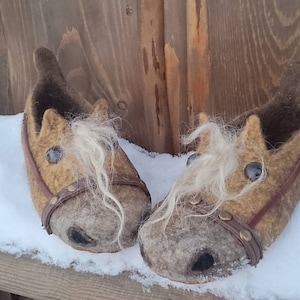 May include: A pair of brown felt horse head slippers with a brown leather strap and white felt mane. The slippers are sitting on a wooden surface covered in snow.