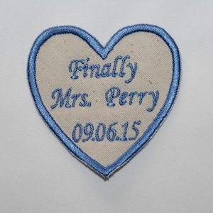 May include: A white heart-shaped patch with blue stitching around the edges. The patch has the text "Finally Mrs. Perry 09.06.15" embroidered in white thread.