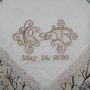 Puede incluir: Pañuelo blanco bordado con un monograma dorado "C&F" y la fecha "May 14, 2016". El pañuelo tiene un borde de encaje blanco.