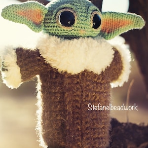 Op de afbeelding: Een gehaakt groen en bruin pluche speelgoed met grote ogen en een witte bontkraag. Het speelgoed zit op een tak. De tekst "Stefanelbeadwork" is zichtbaar in de rechterbenedenhoek van de afbeelding.