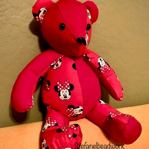 Op de afbeelding: Een rode pluche teddybeer met een Minnie Mouse-ontwerp. De beer zit op een lichtoranje bank. De beer heeft zwarte knoopjesogen en een zwarte neus.