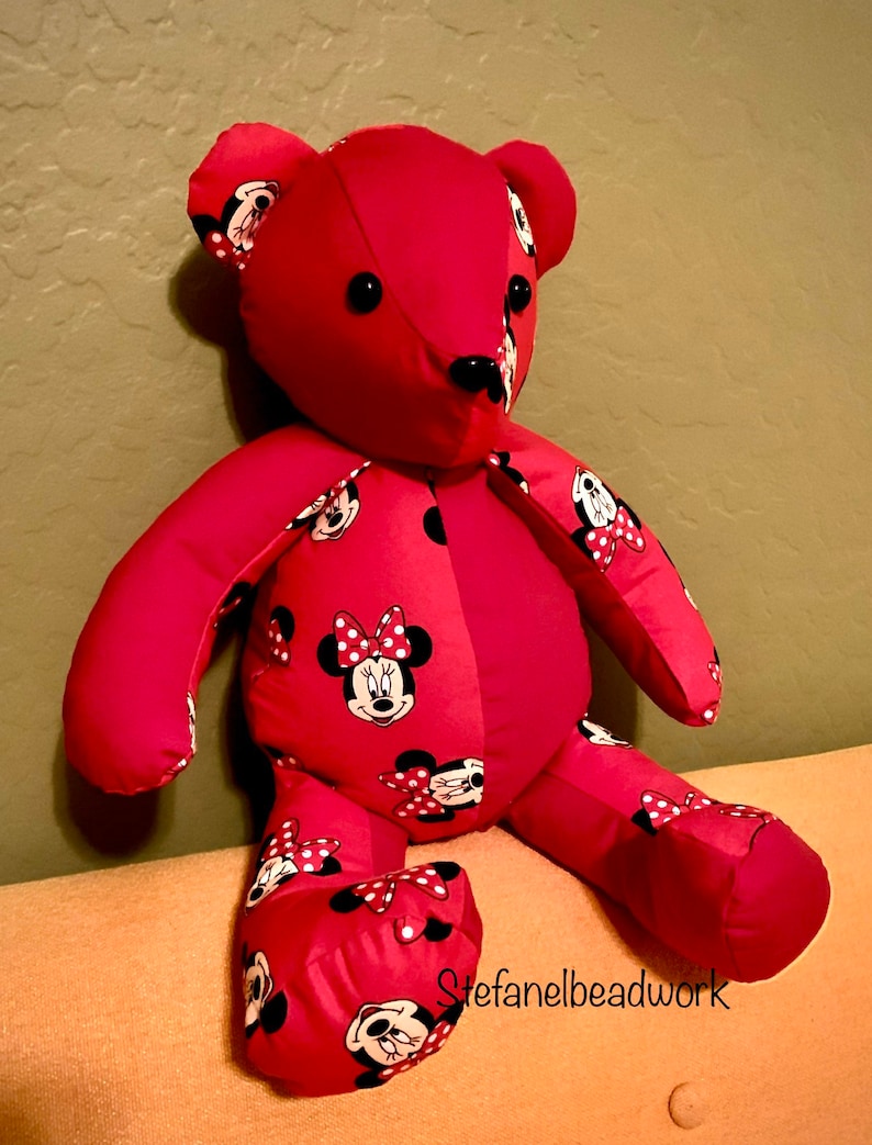 Op de afbeelding: Een rode pluche teddybeer met een Minnie Mouse-ontwerp. De beer zit op een lichtoranje bank. De beer heeft zwarte knoopjesogen en een zwarte neus.