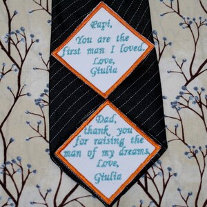 May include: Two white diamond-shaped patches with orange borders and blue text sewn onto a black striped tie. The top patch reads "Papi, You are the first man I loved. Love, Giulia". The bottom patch reads "Dad, thank you for raising the man of my dreams. Love, Giulia".