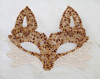 May include: A delicate lace mask with brown and white detailing, resembling a fox face. The mask has two long ties for attaching to the head.