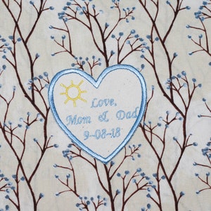 Può includere: Un cerotto a forma di cuore blu con un sole e il testo "Love, Mom & Dad 9-08-18" su uno sfondo in tessuto beige con un motivo floreale marrone.
