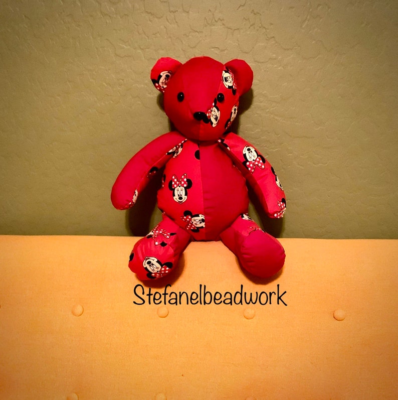 Op de afbeelding: Een rode teddybeer van stof met een Mickey Mouse-print. De beer zit op een lichtoranje bank met een knoopsteekdesign. De tekst "stefanelbeadwork" is zichtbaar in het midden onderaan de afbeelding.