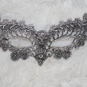 May include: A silver lace masquerade mask with a delicate floral design. The mask has a black ribbon attached to the sides.