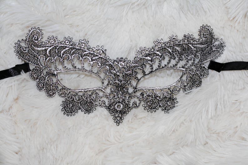 May include: A silver lace masquerade mask with a delicate floral design. The mask has a black ribbon attached to the sides.