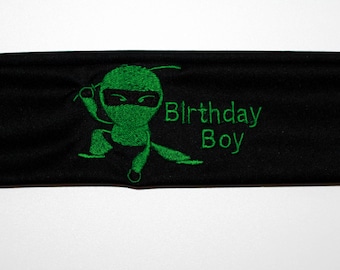 Puede incluir: Cinta para la cabeza negra con un diseño de ninja bordado en verde y la frase "Birthday Boy". El ninja está representado en una postura de lucha. La cinta está hecha de un material suave y elástico.