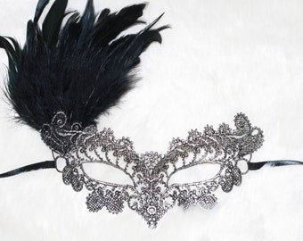 Può includere: Una maschera da ballo in pizzo nero con una piuma nera attaccata. La maschera ha un design delicato ed è decorata con strass.