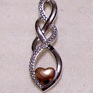 May include: Silver-tone pendant with an infinity design, accented with clear crystals and a gold-tone heart at the bottom. The pendant is likely made of metal and is designed to be worn as jewelry.