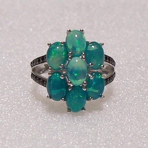 May include: A silver ring featuring a cluster of seven oval-shaped, iridescent green opals arranged in a floral pattern. The band is split and accented with small, dark, round stones along the sides, creating a contrast with the bright opals.