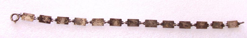 Peut inclure: Un bracelet cha&icirc;ne argent&eacute; avec des maillons rectangulaires. Le bracelet est dot&eacute; d'un petit fermoir &agrave; une extr&eacute;mit&eacute; et est pr&eacute;sent&eacute; sur un fond blanc. Les maillons semblent ternis, ce qui lui donne un aspect vintage.