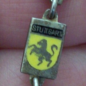 Peut inclure: Un bracelet de charme vintage en argent avec des breloques rectangulaires affichant des armoiries de villes. Les breloques incluent Hambourg, Stuttgart et Munich. Le bracelet est reli&eacute; par de petits anneaux argent&eacute;s.