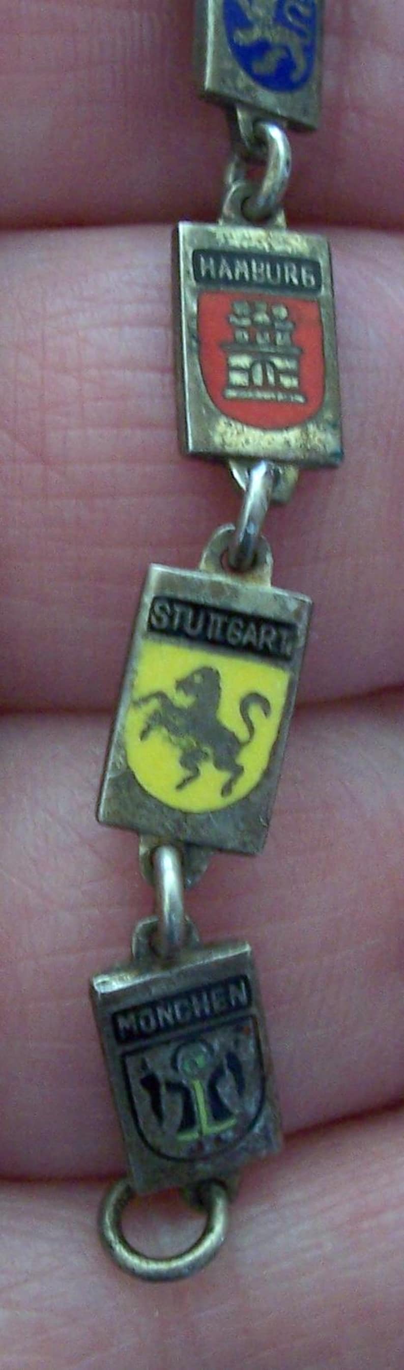 Peut inclure: Un bracelet de charme vintage en argent avec des breloques rectangulaires affichant des armoiries de villes. Les breloques incluent Hambourg, Stuttgart et Munich. Le bracelet est reli&eacute; par de petits anneaux argent&eacute;s.