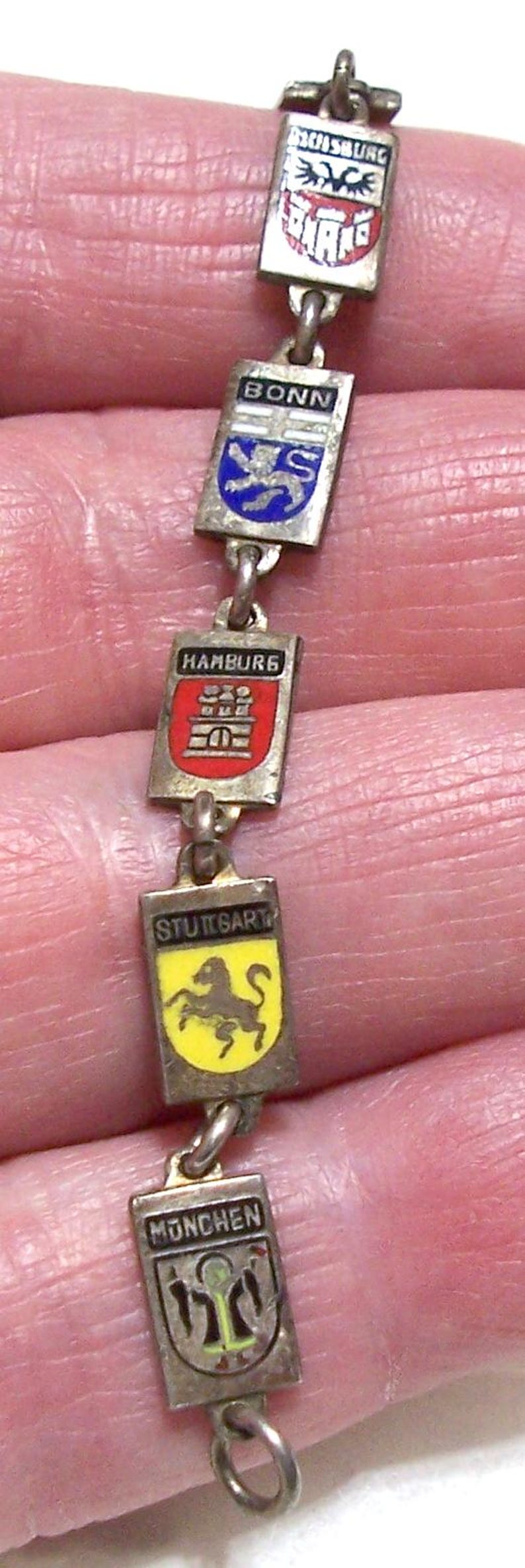 Peut inclure: Bracelet argent&eacute; compos&eacute; de breloques rectangulaires avec des armoiries &eacute;maill&eacute;es. Les breloques affichent les noms et les embl&egrave;mes des villes allemandes : Salzbourg, Bonn, Hambourg, Stuttgart et Monchen.