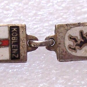 Peut inclure: Un bracelet argent&eacute; orn&eacute; de breloques rectangulaires avec des armoiries de villes en &eacute;mail. Les breloques sont reli&eacute;es par de petits anneaux argent&eacute;s. Les armoiries incluent des noms de villes comme Koblenz, Berlin et Mainz.