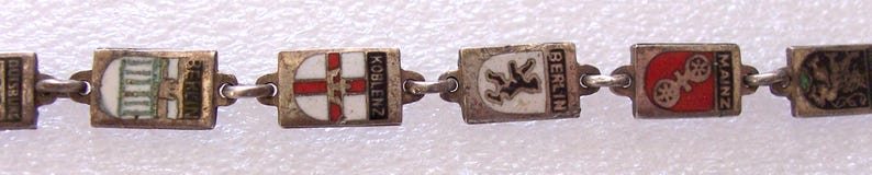Peut inclure: Un bracelet argent&eacute; orn&eacute; de breloques rectangulaires avec des armoiries de villes en &eacute;mail. Les breloques sont reli&eacute;es par de petits anneaux argent&eacute;s. Les armoiries incluent des noms de villes comme Koblenz, Berlin et Mainz.