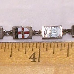 Peut inclure: Un bracelet vintage avec des breloques rectangulaires, chacune orn&eacute;e d'un embl&egrave;me color&eacute; diff&eacute;rent. Le bracelet mesure environ 18 cm de long et poss&egrave;de une cha&icirc;ne et un fermoir en m&eacute;tal argent&eacute;. Les breloques pr&eacute;sentent divers motifs, dont des armoiries et des symboles h&eacute;raldiques.
