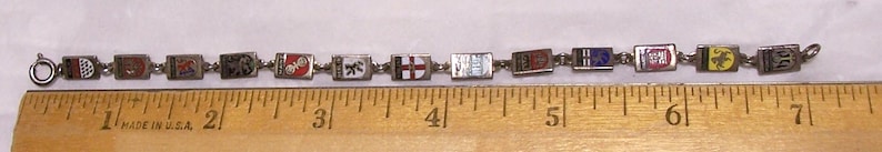 Peut inclure: Un bracelet vintage avec des breloques rectangulaires, chacune orn&eacute;e d'un embl&egrave;me color&eacute; diff&eacute;rent. Le bracelet mesure environ 18 cm de long et poss&egrave;de une cha&icirc;ne et un fermoir en m&eacute;tal argent&eacute;. Les breloques pr&eacute;sentent divers motifs, dont des armoiries et des symboles h&eacute;raldiques.