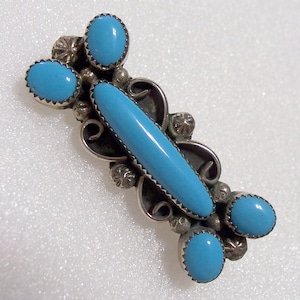 May include: A silver-toned brooch featuring a central, elongated turquoise stone, flanked by four oval turquoise stones. The brooch has a detailed, ornate design with decorative silver accents and a pin closure on the back. The turquoise stones are a bright, vibrant blue.