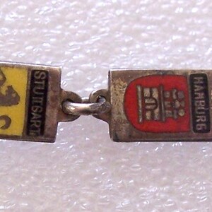 Peut inclure: Un bracelet vintage en argent avec des breloques rectangulaires orn&eacute;es d'&eacute;cussons de villes &eacute;maill&eacute;s color&eacute;s. Les breloques sont reli&eacute;es par de petits anneaux en argent. Les villes incluent Munich, Stuttgart, Hambourg, Bonn, et d'autres.