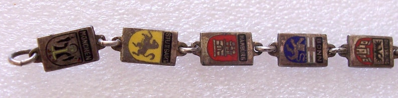 Peut inclure: Un bracelet vintage en argent avec des breloques rectangulaires orn&eacute;es d'&eacute;cussons de villes &eacute;maill&eacute;s color&eacute;s. Les breloques sont reli&eacute;es par de petits anneaux en argent. Les villes incluent Munich, Stuttgart, Hambourg, Bonn, et d'autres.