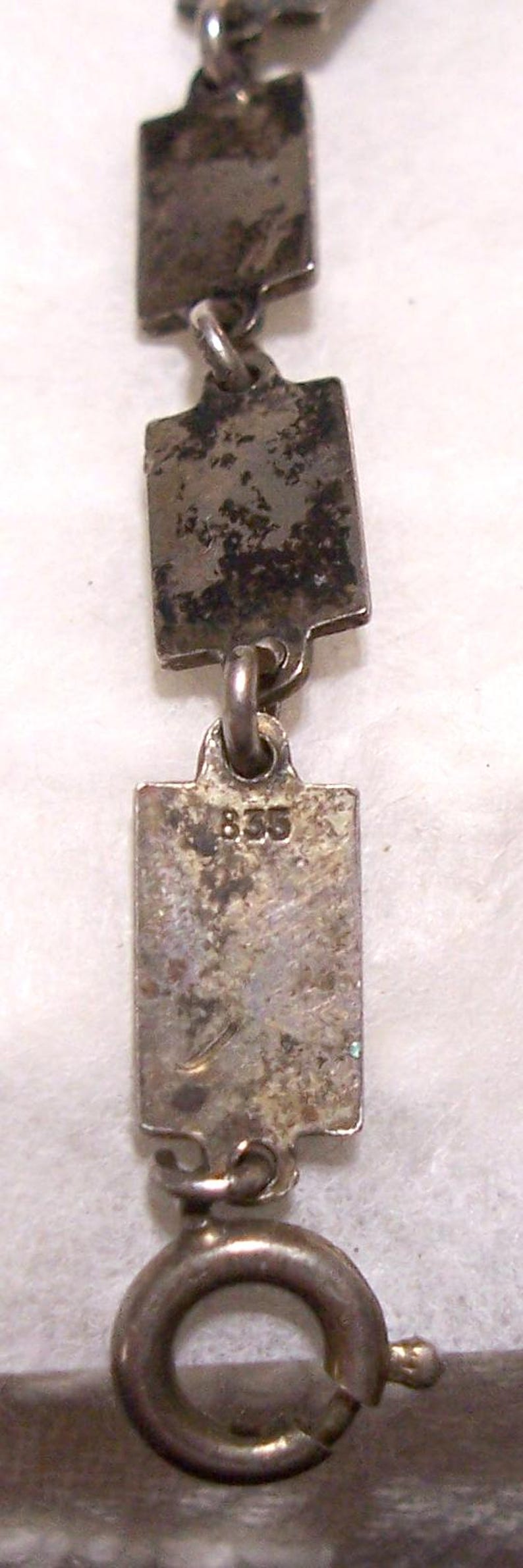 Peut inclure: Gros plan sur un collier argent&eacute; avec des maillons rectangulaires. Les maillons sont reli&eacute;s par de petits anneaux. Le fermoir est un anneau &agrave; ressort circulaire. Le chiffre "835" est visible sur l'un des maillons.