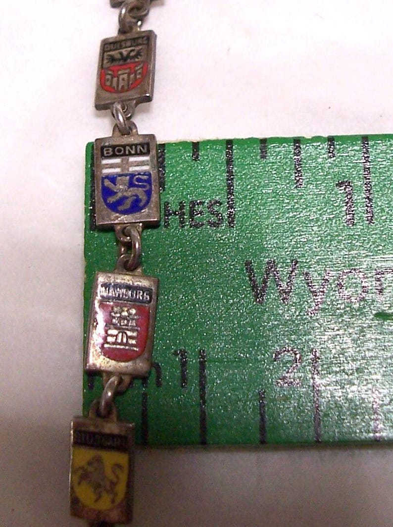 Peut inclure: Un collier en argent avec des pendentifs rectangulaires orn&eacute;s d'armoiries de villes. Les armoiries incluent les noms de villes allemandes comme Bonn et Stuttgart. Une r&egrave;gle verte est en arri&egrave;re-plan.