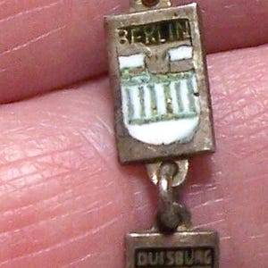 Peut inclure: Un bracelet de charme vintage en argent avec des breloques rectangulaires &eacute;maill&eacute;es. Les breloques affichent des noms de villes comme Berlin, Koblenz et Bonn. Le bracelet est tenu par des doigts.