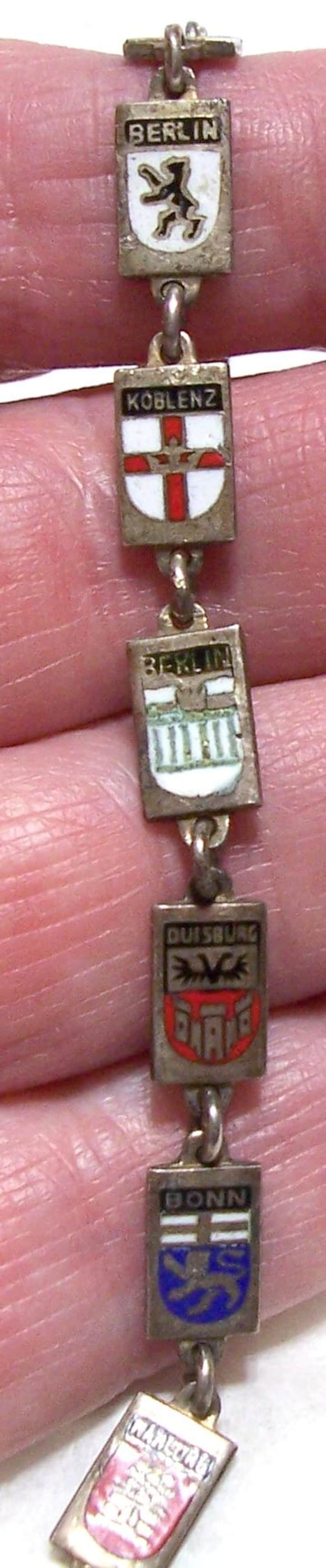 Peut inclure: Un bracelet de charme vintage en argent avec des breloques rectangulaires &eacute;maill&eacute;es. Les breloques affichent des noms de villes comme Berlin, Koblenz et Bonn. Le bracelet est tenu par des doigts.