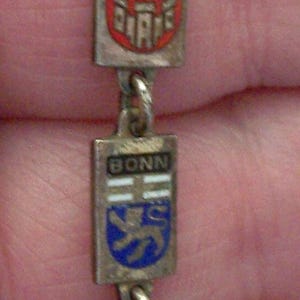 Peut inclure: Un bracelet argent&eacute; orn&eacute; de breloques rectangulaires avec des motifs &eacute;maill&eacute;s. Les breloques affichent des noms de villes comme D&uuml;sseldorf, Bonn et Hambourg, chacun avec un embl&egrave;me unique. Le bracelet est reli&eacute; par de petits anneaux m&eacute;talliques.