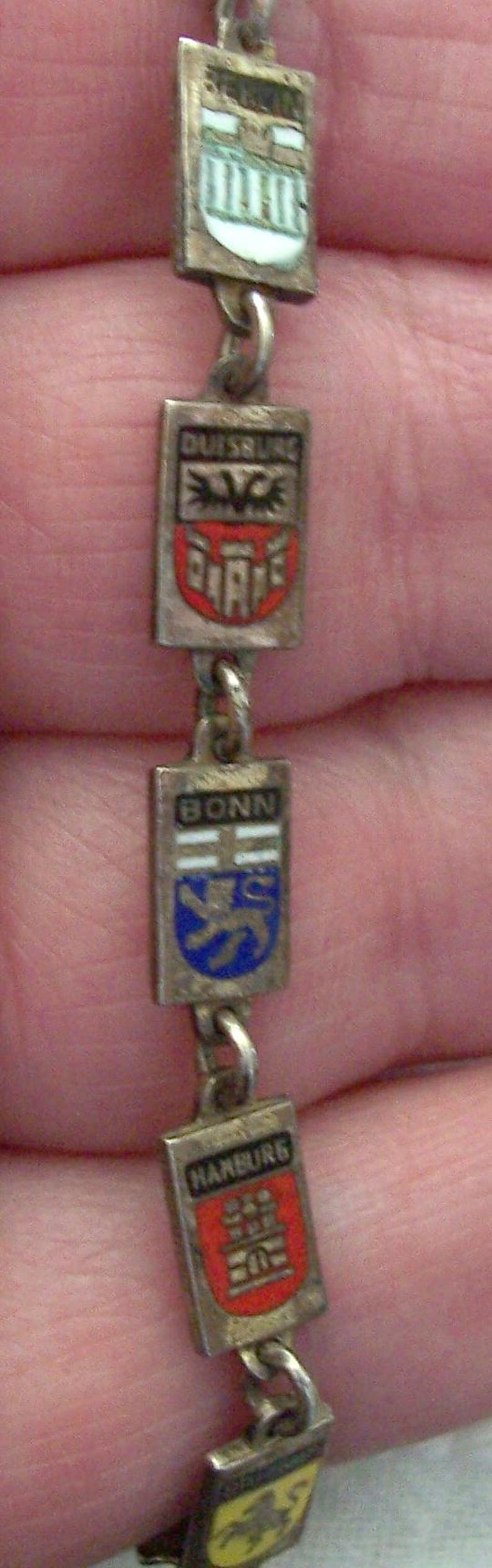 Peut inclure: Un bracelet argent&eacute; orn&eacute; de breloques rectangulaires avec des motifs &eacute;maill&eacute;s. Les breloques affichent des noms de villes comme D&uuml;sseldorf, Bonn et Hambourg, chacun avec un embl&egrave;me unique. Le bracelet est reli&eacute; par de petits anneaux m&eacute;talliques.