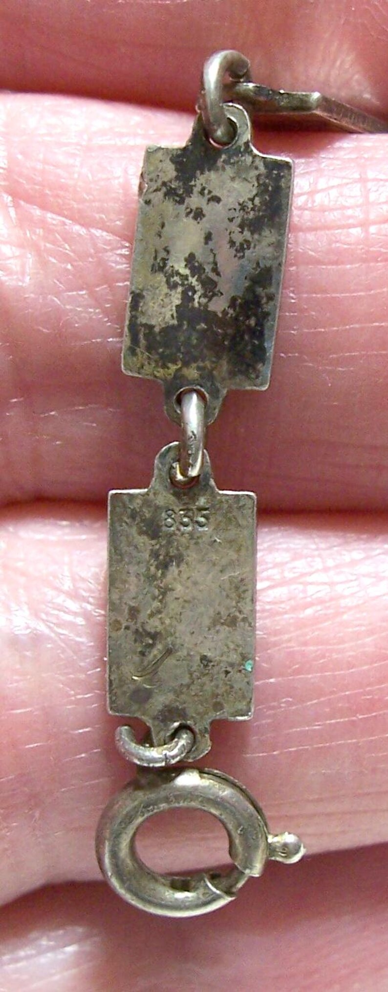 Peut inclure: Gros plan sur un collier en argent avec des maillons rectangulaires. Les maillons sont ternis, montrant des signes d'&acirc;ge. Un maillon est estampill&eacute; du num&eacute;ro "835". Le collier a un fermoir &agrave; ressort.