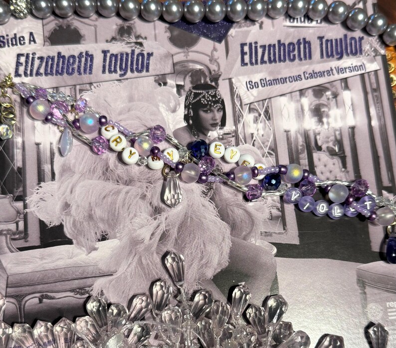Puede incluir: Un collar de cuentas con cuentas moradas, blancas e iridiscentes que deletrean "CRY" y "VIOLET" se muestra sobre una imagen en blanco y negro de Elizabeth Taylor. La imagen es de una portada de &aacute;lbum. Tambi&eacute;n se ve un collar de perlas y detalles de cristal.