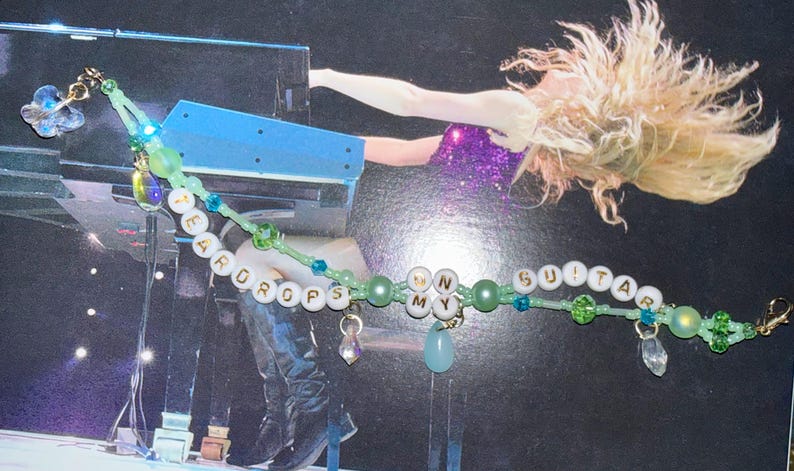 K&ouml;nnte beinhalten: Ein Perlenarmband mit den W&ouml;rtern "TEARDROPS" und "GUITAR" in wei&szlig;en Buchstabenperlen. Das Armband hat gr&uuml;ne und blaue Perlen, sowie h&auml;ngende Charms und einen goldenen Verschluss. Die Buchstaben "NY" sind ebenfalls enthalten.