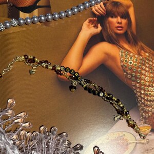 Puede incluir: Una pulsera dorada con cuentas negras que deletrean la palabra "FATHER" y peque&ntilde;os dijes de coraz&oacute;n. La pulsera est&aacute; adornada con cuentas burdeos y doradas. Tambi&eacute;n se ve un collar de perlas plateadas.