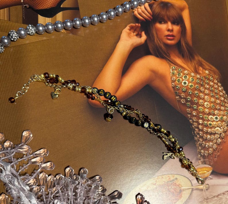 Puede incluir: Una pulsera dorada con cuentas negras que deletrean la palabra "FATHER" y peque&ntilde;os dijes de coraz&oacute;n. La pulsera est&aacute; adornada con cuentas burdeos y doradas. Tambi&eacute;n se ve un collar de perlas plateadas.