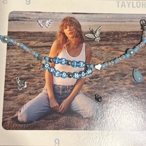 May include: A beaded necklace with the words "is it over now" in blue and silver beads. The necklace has light blue and white beads, a heart charm, and a teardrop pendant. Additional charms include a butterfly, a bird, and a lock.