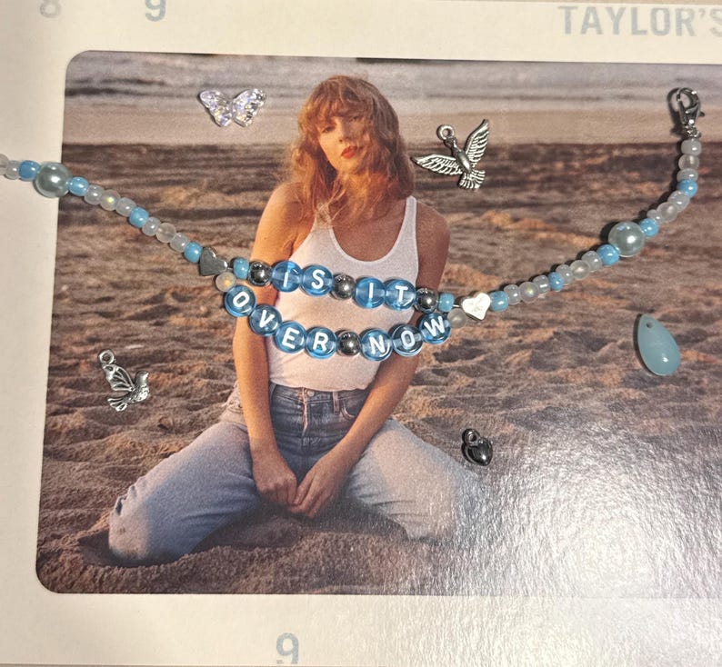 May include: A beaded necklace with the words "is it over now" in blue and silver beads. The necklace has light blue and white beads, a heart charm, and a teardrop pendant. Additional charms include a butterfly, a bird, and a lock.