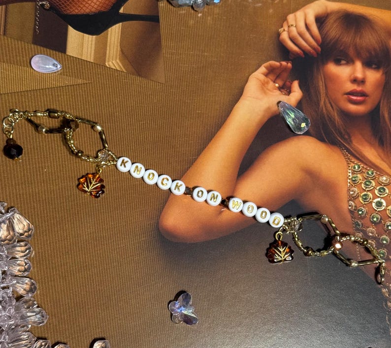 Puede incluir: Una pulsera dorada con las palabras "KNOCK ON WOOD" escritas con cuentas blancas. La pulsera presenta peque&ntilde;os dijes y est&aacute; colocada sobre un fondo marr&oacute;n con una mujer en el fondo.