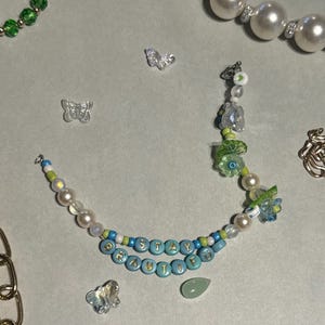 K&ouml;nnte beinhalten: Eine Halskette mit den Worten "STAY BEAUTIFUL" in blauen Perlen. Die Halskette hat Perlen, gr&uuml;ne und blaue Perlen und Schmetterlingsanh&auml;nger. Weitere Schmuckst&uuml;cke, darunter eine Perlenkette und eine goldene Kette, sind ebenfalls sichtbar.
