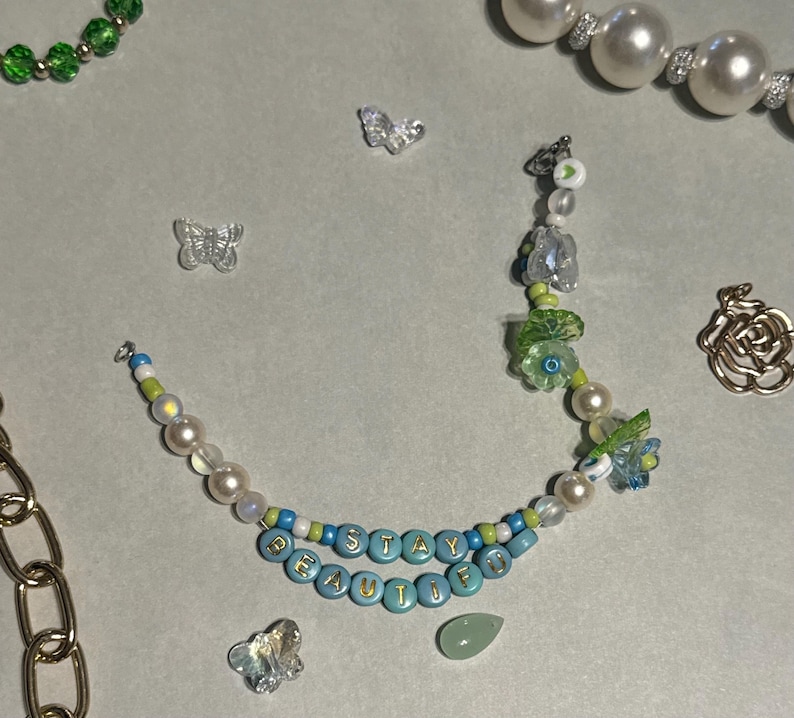K&ouml;nnte beinhalten: Eine Halskette mit den Worten "STAY BEAUTIFUL" in blauen Perlen. Die Halskette hat Perlen, gr&uuml;ne und blaue Perlen und Schmetterlingsanh&auml;nger. Weitere Schmuckst&uuml;cke, darunter eine Perlenkette und eine goldene Kette, sind ebenfalls sichtbar.