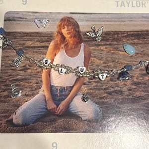 May include: A silver beaded bracelet with the letters "S L U T" spelled out in white heart-shaped beads. The bracelet also features charms of birds, butterflies, and a heart. The background shows a person sitting on a beach.