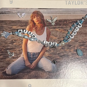 May include: A beaded bracelet with the words "WISH YOU COULD" in white letter beads, accented with blue beads and charms. The bracelet is displayed on a photograph with a beach scene and a person wearing a white tank top and jeans.